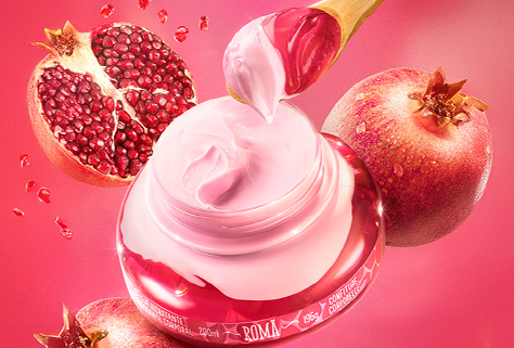 A imagem mostra um frasco de creme com um tom rosado, com uma colher de madeira retirando o produto, ao lado de romãs ao fundo. O cenário é vibrante, com uma tonalidade de rosa predominante, criando um visual suave e delicado.