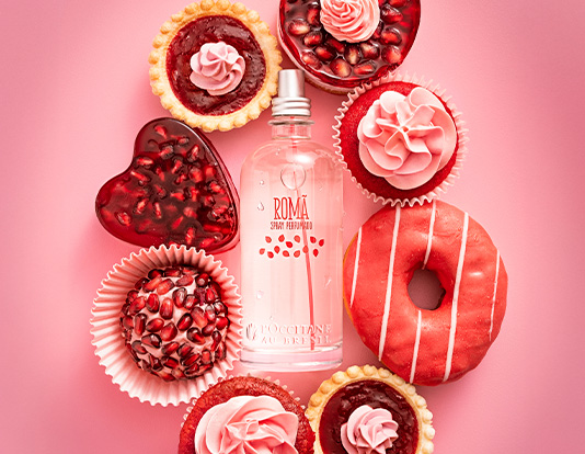 A imagem mostra um frasco de perfume no centro, cercado por doces decorados em tons de rosa e vermelho sobre um fundo rosa. Entre os doces, há cupcakes, tortinhas, um donut e um bombom em forma de coração.