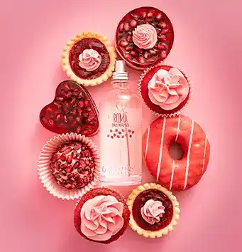 A imagem mostra um frasco de perfume no centro, cercado por doces decorados em tons de rosa e vermelho sobre um fundo rosa. Entre os doces, há cupcakes, tortinhas, um donut e um bombom em forma de coração.